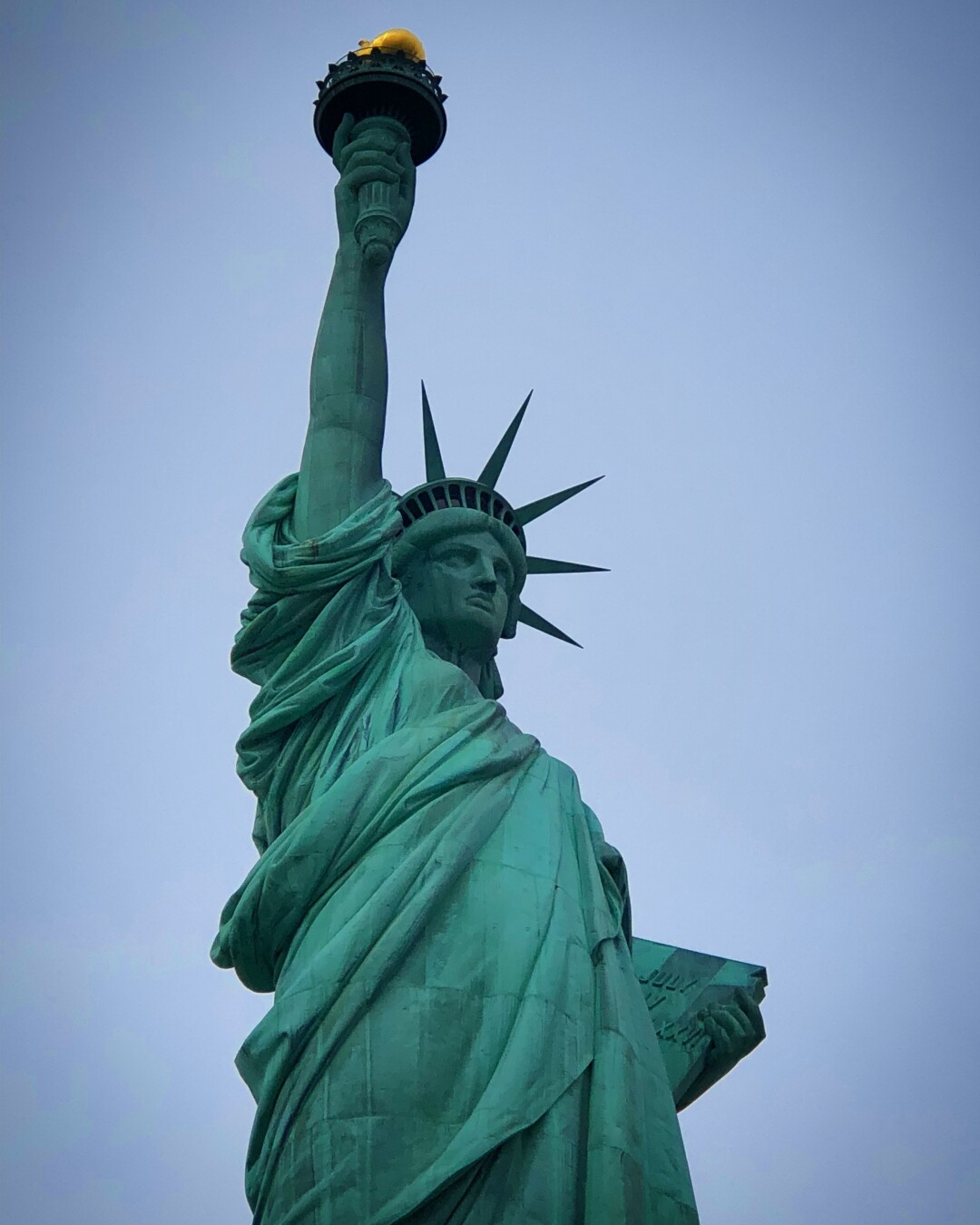 The image features the Statue of Liberty, a towering green statue with a raised torch and a book in her left hand, set against a pale blue sky. The statue symbolizes freedom and democracy.