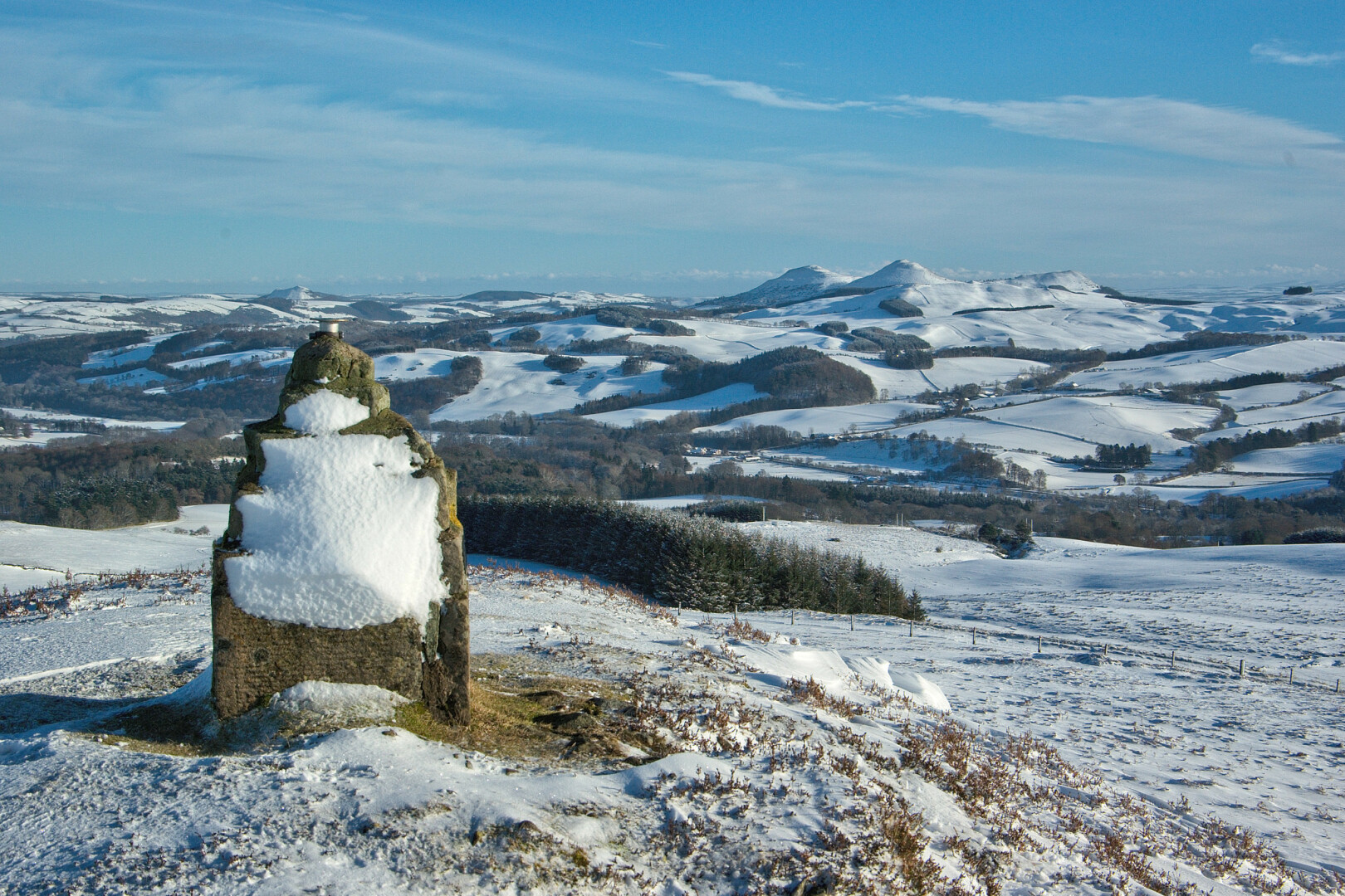 A view east from the summit of Linglie Hill, near Selkirk, to the Eildon Hills, near Melrose, 9 km distant, over a snow-covered landscape. Linglie Hill's rectangular, stepped stone cairn is in the left foreground, plastered with snow on its western side; it has a metal mount at the top, capping an opening in which a UK Union Flag is placed on 'Common Riding' day in June. On the right skyline are the three Eildon Hills. The land between is a mixture of fields and trees, some in (mainly small-scale) coniferous plantations; it's a sunny day, with some high wispy cloud, but plenty of blue sky, and very clear air.