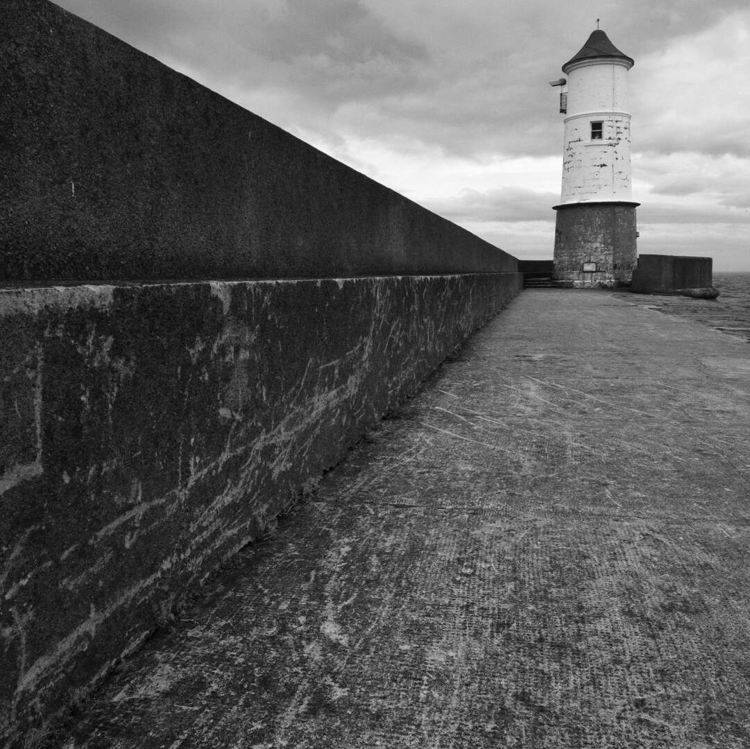 A wide-angle black-and-white shot of the lighthouse at the end of the pier, taken from the pier, at Berwick-upon-Tweed. The lighthouse, a circular structure with a dark conical roof, a white mid-section and a dark stone base, is in the top right hand corner of the image. The dark concreted sea wall, stepped at half-height, initially occupies the whole of the left side of the image, but the effect of it apparently getting smaller as the distance away from the camera increases, makes it seem like an arow head pointing to the base of the lighthouse. There's a similar effect with the lighter grey concreted pier walkway, which initially occupies the whole of the bottom of the image. It's a dull day, with thick grey clouds visible behind the lighthouse, and just a glimpse of the sea.