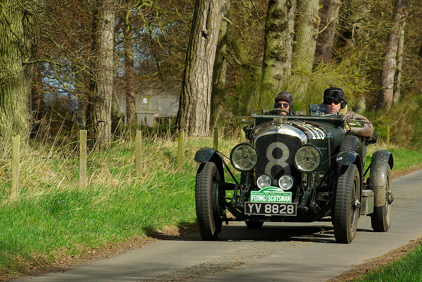 A medium telephoto shot of a 'vintage' open-top car, a 1928 4.5 litre Bentley, driving down a single-track country road at a moderate speed. It is viewed mainly front-on, but the nearside is visible; it is painted black, with some chrome parts. It has two occupants, wearing thick clothing and lined leather hats. The left side of the bonnet has the number 115 painted on it in white, and the radiator grill displays a large silver number 8; the car's registration number is YV 8828, above which is a plaque indicating it is taking part in the 'Flying Scotsman 2015' rally, a 'Vintage Reliability Trial'. The front of the car sports two large main headlamps, and two smaller spotlights. On the other side of the road is a grass verge, a wooden post-and-wire fence, and a narrow stand of leafless trees, through a gap in which part of a farm building can be seen. It's a bright sunny day. the shadow of the car falls to its right.