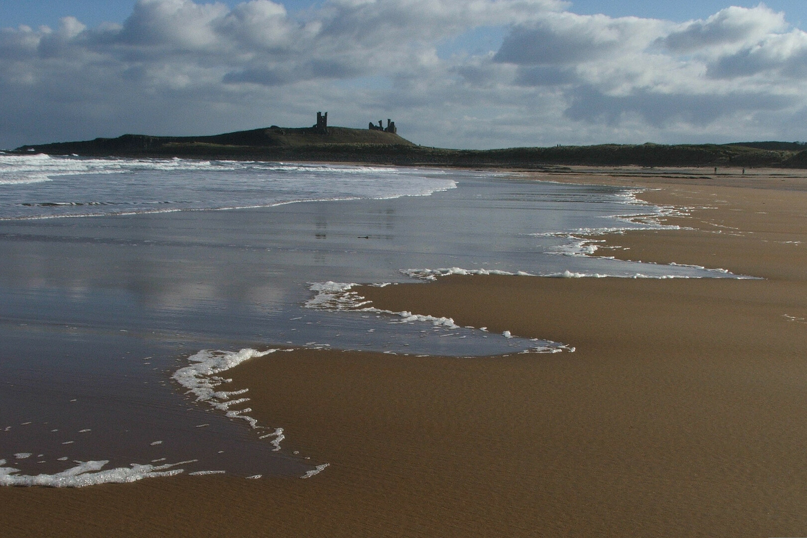 A wide-angle view south across Embleton Bay to the ruins of Dunstanburgh Castle. The foreground and righthand side of the image shows the serrated edges of a wet sandy beach as the tide has receded, flecked with foam; the left side the receding sea. The top third of the image shows land jutting out into the sea, with the ruins of the castle on the left skyline. The sky behind is mainly cloudy, with the some patches of blue sky.