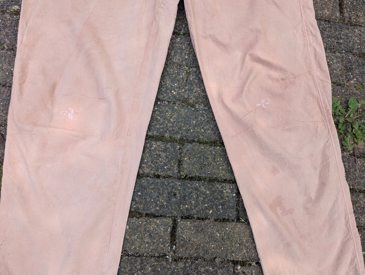 Close-up of the front knees on a pair of tan cotton pants, featuring small decorative or reinforcing stitched cross-shaped designs over the knee darts.