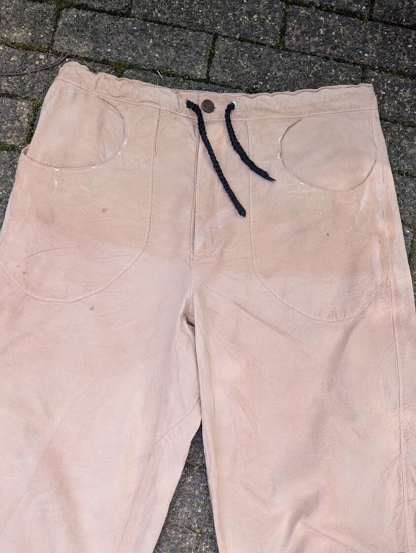 Close-up view of the front waistband of a pair of tan cotton pants, featuring a dark black braided drawstring and curved pocket openings.