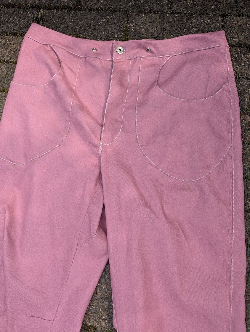 Close-up view of the front waistband and curved pockets of a pair of dusty pink denim pants, featuring metal snap fasteners and white topstitching.