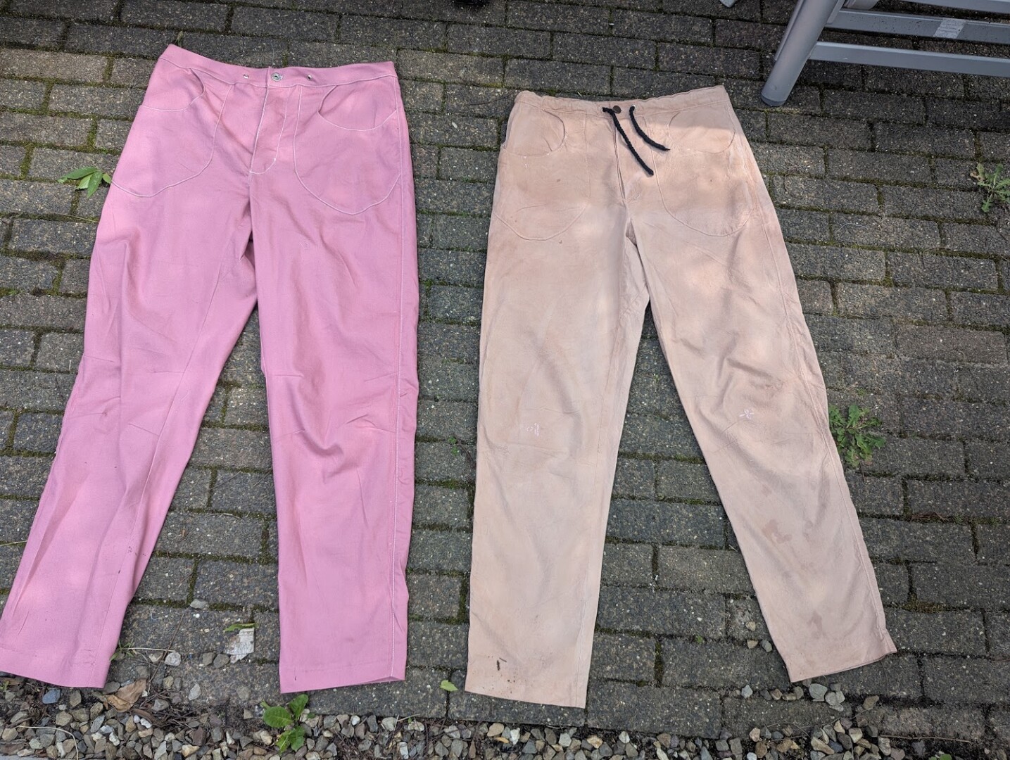 Two pairs of handmade pants laid side-by-side on brick pavers: a dusty pink denim pair on the left and a tan, avocado-dyed cotton pair with a black drawstring on the right.