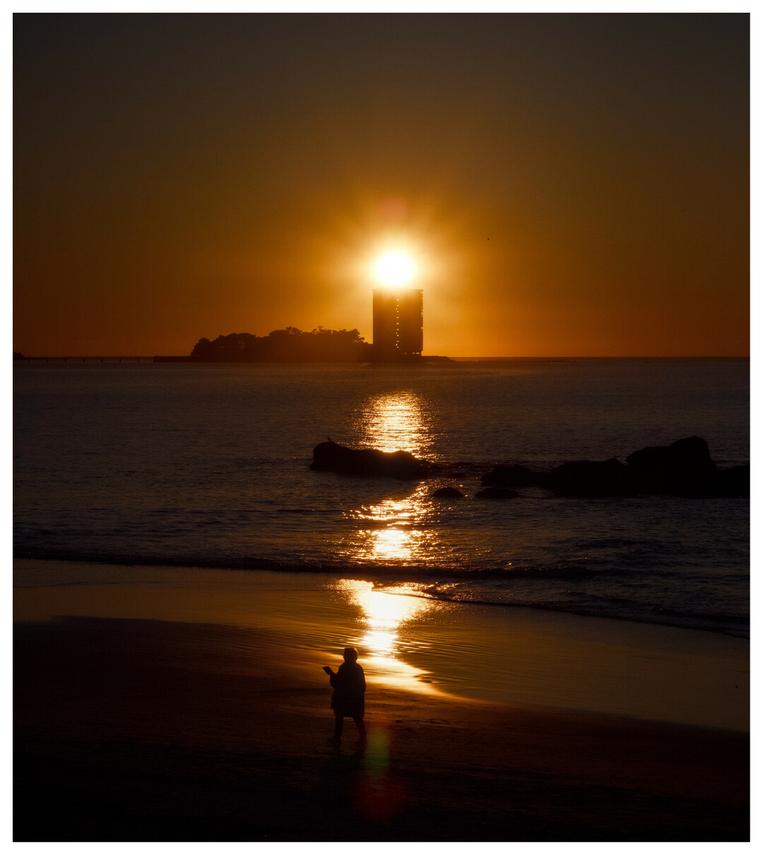 Solpor xusto enriba dun edificio iluminando a praia mentras unha persoa pasea pola praia. (galego).

Sunset just above a building illuminating the beach as a person walks along the beach.