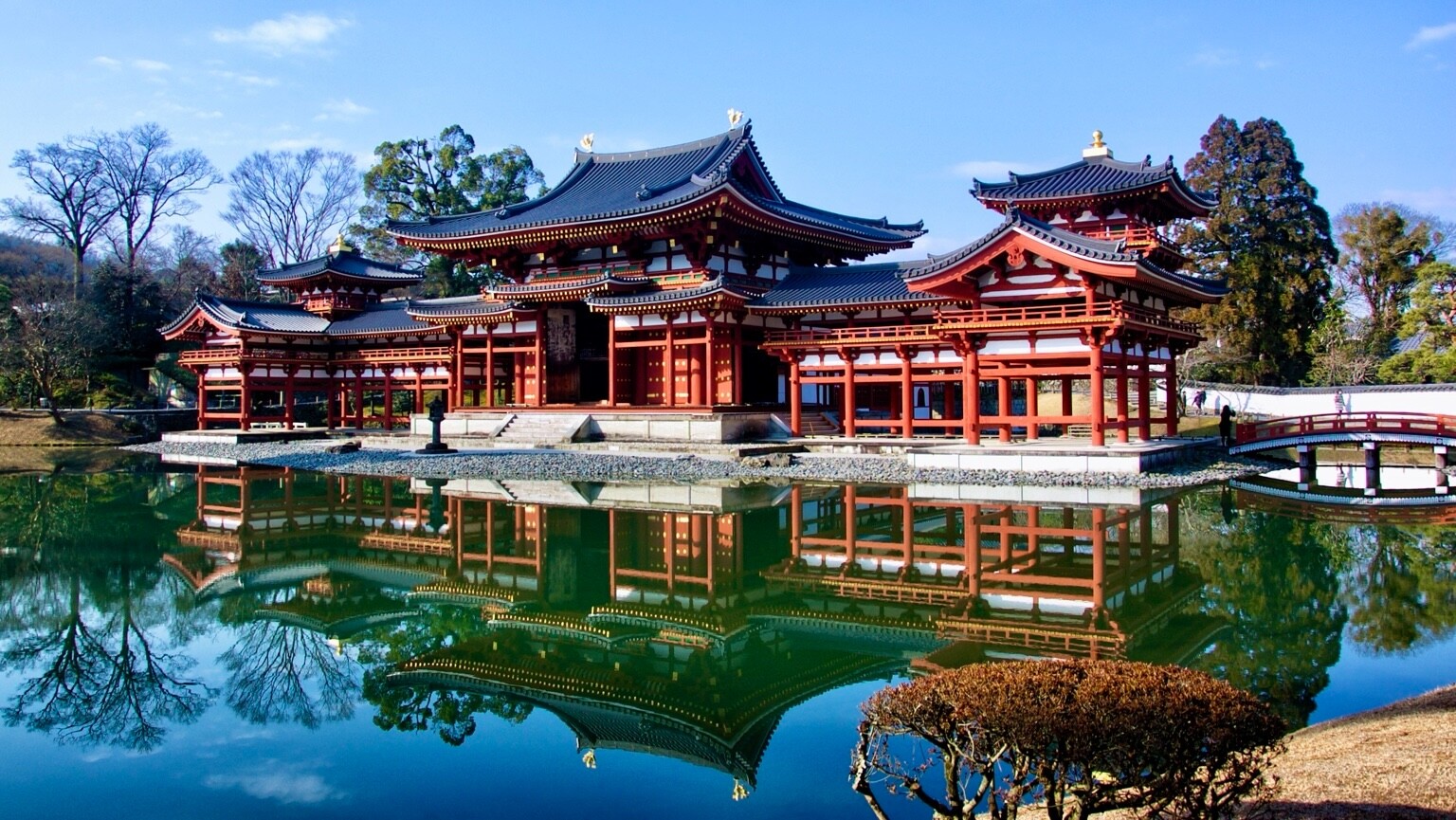 A photo taken of a Buddhist temple in the city of Uji in Kyoto Prefecture.
The wood pillars are all painted red. In front of the temple is a lake, appearing blue in the photo. Behind the temple are trees ans the sky is in a lighter shade of blue then the water.