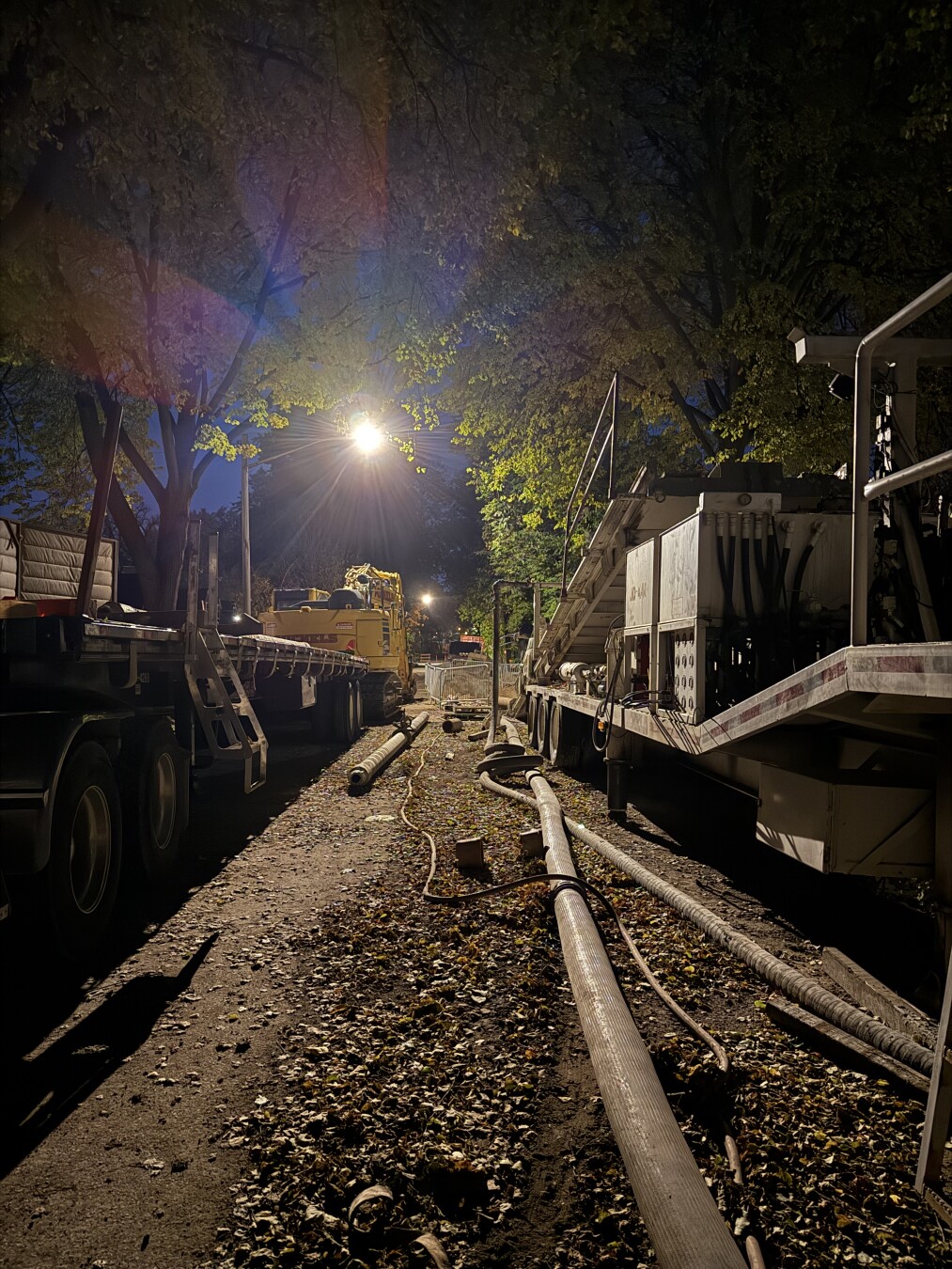 photo of a residential street at night, both sides lined with construction equipment, with a harsh streetlight in the upper center