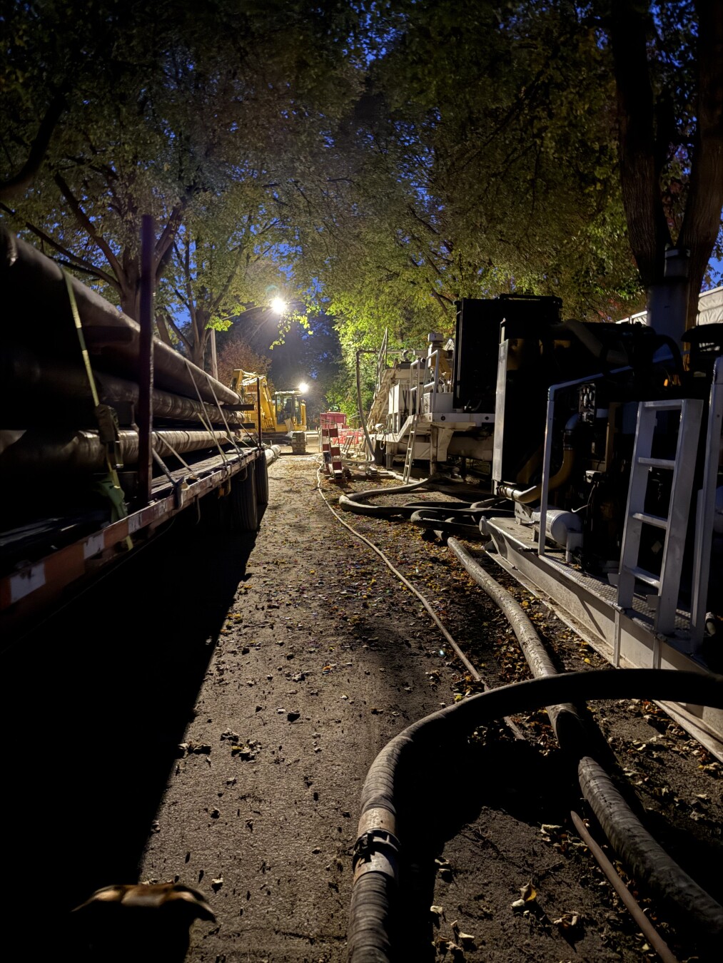photo of a residential street at night, both sides lined with construction equipment, with a harsh streetlight in the upper center