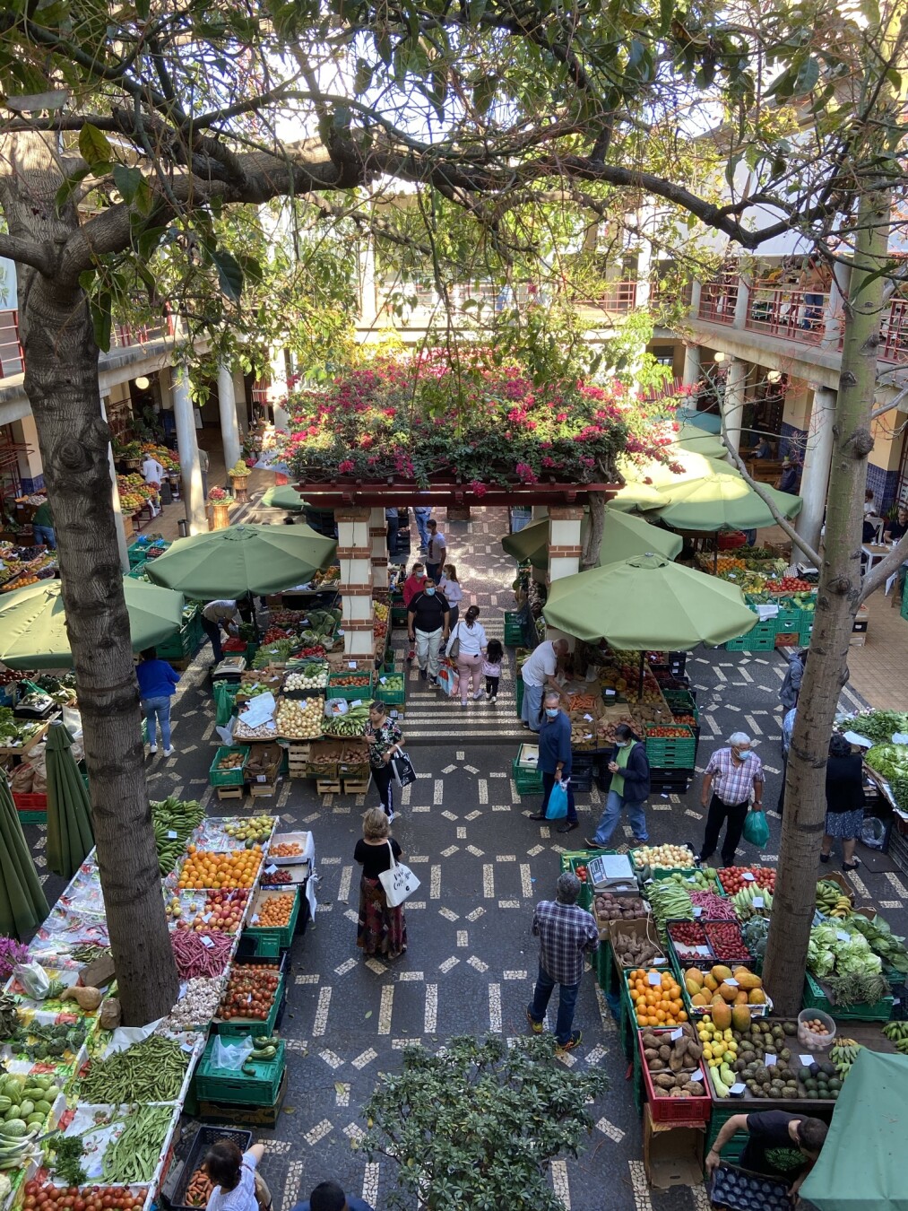 Das Bild zeigt einen belebten Markt mit frischem Obst und Gemüse, aufgenommen von einer erhöhten Perspektive. Die Marktstände sind mit grünen Sonnenschirmen bedeckt und präsentieren eine bunte Vielfalt an Produkten wie Tomaten, Orangen, Bananen, Bohnen und Knoblauch. Das Pflaster des Marktes hat ein kunstvolles Muster aus dunklen und hellen Steinen.

In der Mitte des Bildes befindet sich ein überdachter Eingang mit roten und weißen Zierelementen, der mit blühenden Pflanzen bewachsen ist. Menschen bewegen sich durch den Markt, einige tragen Einkaufstaschen, andere unterhalten sich oder begutachten die Waren. Die Architektur des Marktes hat eine offene, luftige Gestaltung mit umlaufenden Balkonen im oberen Bereich, von denen aus weitere Besucher auf das Marktgeschehen blicken können.