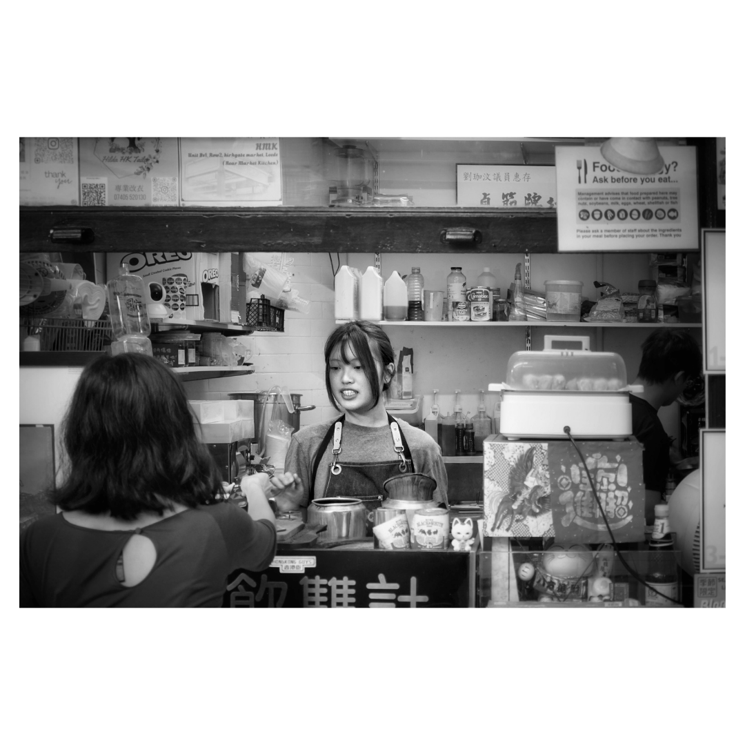 This is a landscape format black and white image of a young woman serving a customer through the hatch of a Chinese street food stall in Leeds market.