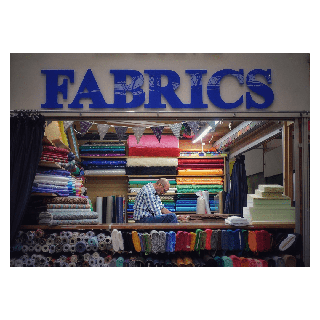 This is a landscape format colour image of a man sitting within a fabric stall within an indoor market in Leeds. Hello sits hunched over waiting for customers, surrounded by bolts of colourful fabric.