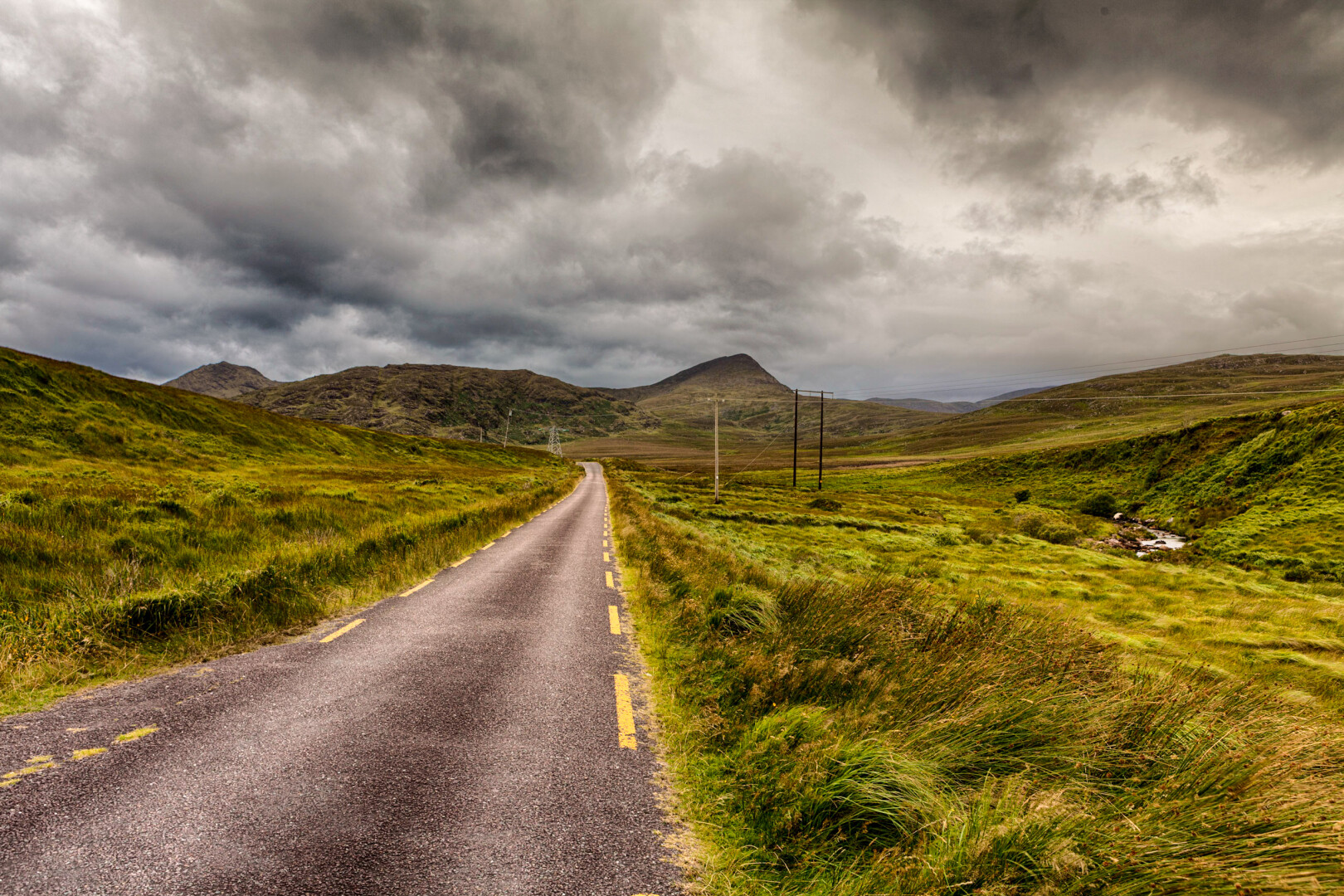 A small road leading into the distance. Mountains in the back. There are dark clouds overhead.