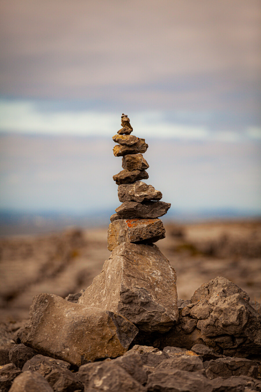 A stack of rocks arranged vertically, set against a blurred natural background. The stones vary in size and shape, creating a balanced tower that rises above larger rocks in the foreground.