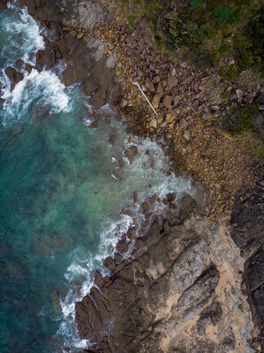 An aerial view of a rocky coastline where waves crash against the shore. The water is a mix of turquoise and clear hues, contrasting with the dark rocks and sandy patches along the beach.