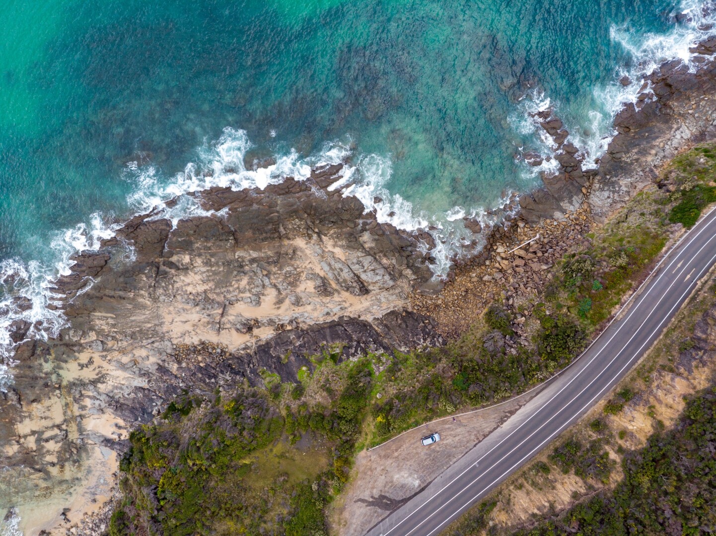 Aerial view of a coastal area featuring rocky shorelines, clear turquoise waters, and a winding road. Waves crash against the rocks, with patches of sandy beach visible. A parked vehicle is seen along the road near the edge of the land.