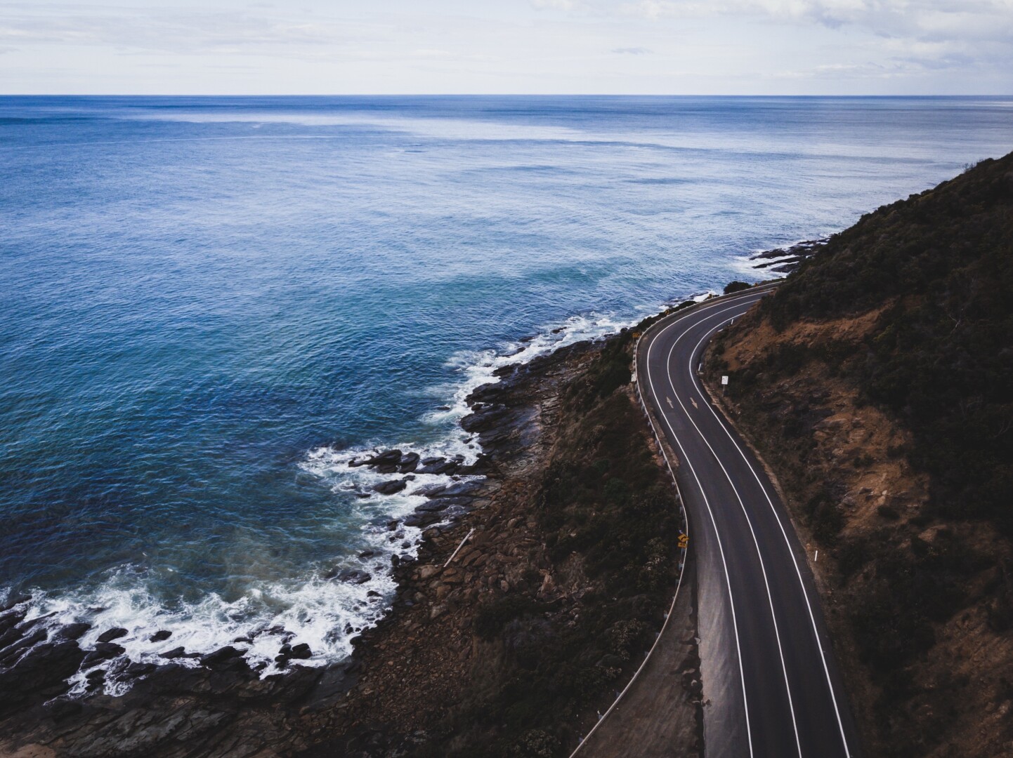 An aerial view of a winding coastal road alongside the ocean. The scene features a calm blue sea with gentle waves lapping against rocky shores and grassy hills. The sky is partly cloudy, creating a serene atmosphere.