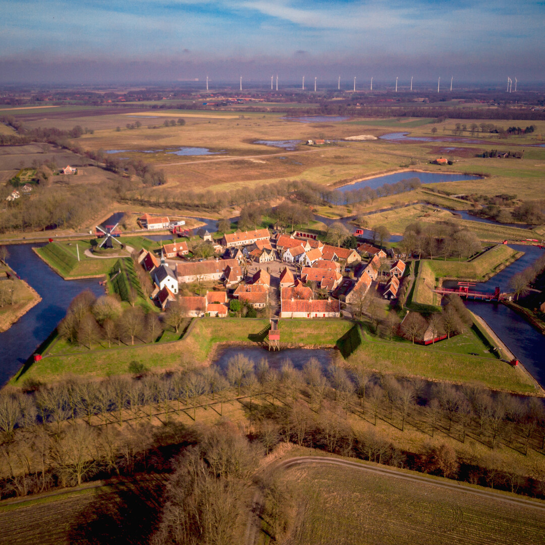 The image is an aerial view of Bourtange, a star-shaped fortress village in the Netherlands. The village is surrounded by a system of moats and green embankments, forming a distinct geometric pattern. The center of the fortress contains red-roofed houses, small streets, and a church, all closely packed together. Several wooden bridges painted red cross over the moats, connecting different sections of the village. Beyond the fortress, the landscape consists of flat fields, patches of trees, and wind turbines in the distance under a clear blue sky.