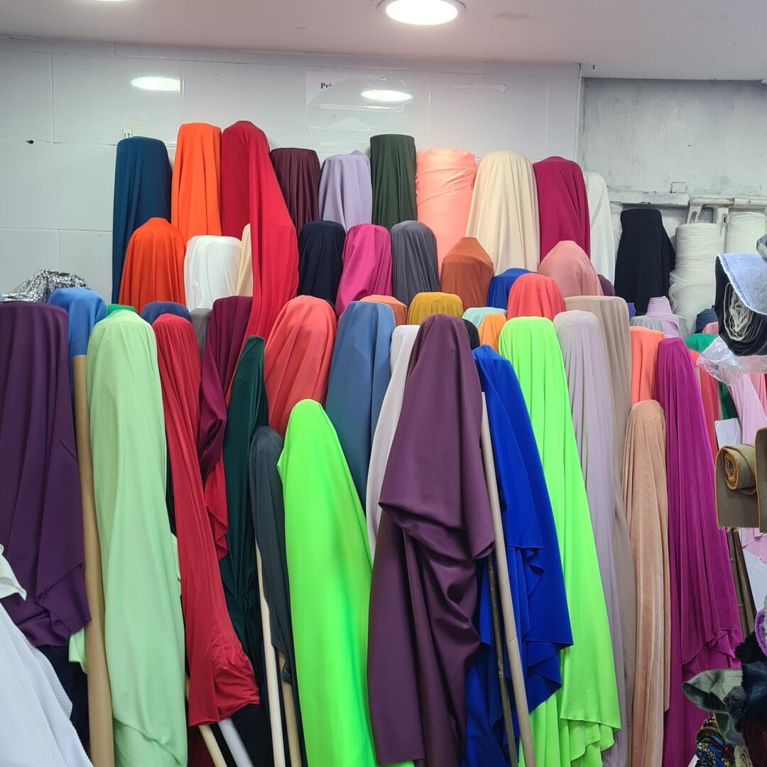thirty or so rolls of fabric stood vertically in three rows. each roll has the end of the fabric draped over it, so they look like very colourful ghosts