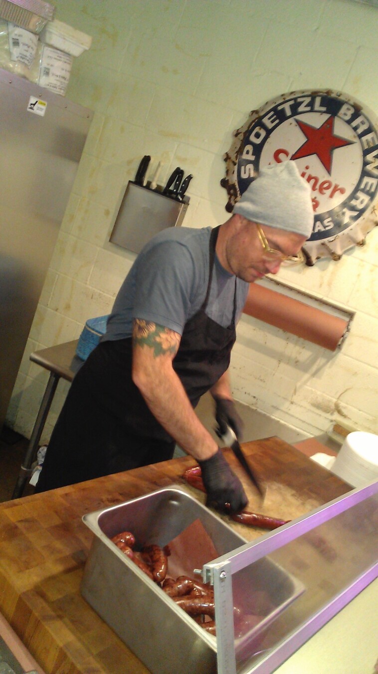 A medium shot of a man (Aaron Franklin) wearing a gray beanie, glasses, a gray t-shirt, and a black apron, standing in a kitchen-like setting. He has a floral tattoo on his left arm. He's wearing black gloves and cutting sausages with a knife on a wooden cutting board. A metal container filled with sausages sits to his left. Behind him, there is a Spoezl Brewery sign on the wall, along with a metal knife holder and packages on a shelf. The walls are light yellow and appear to be tiled.