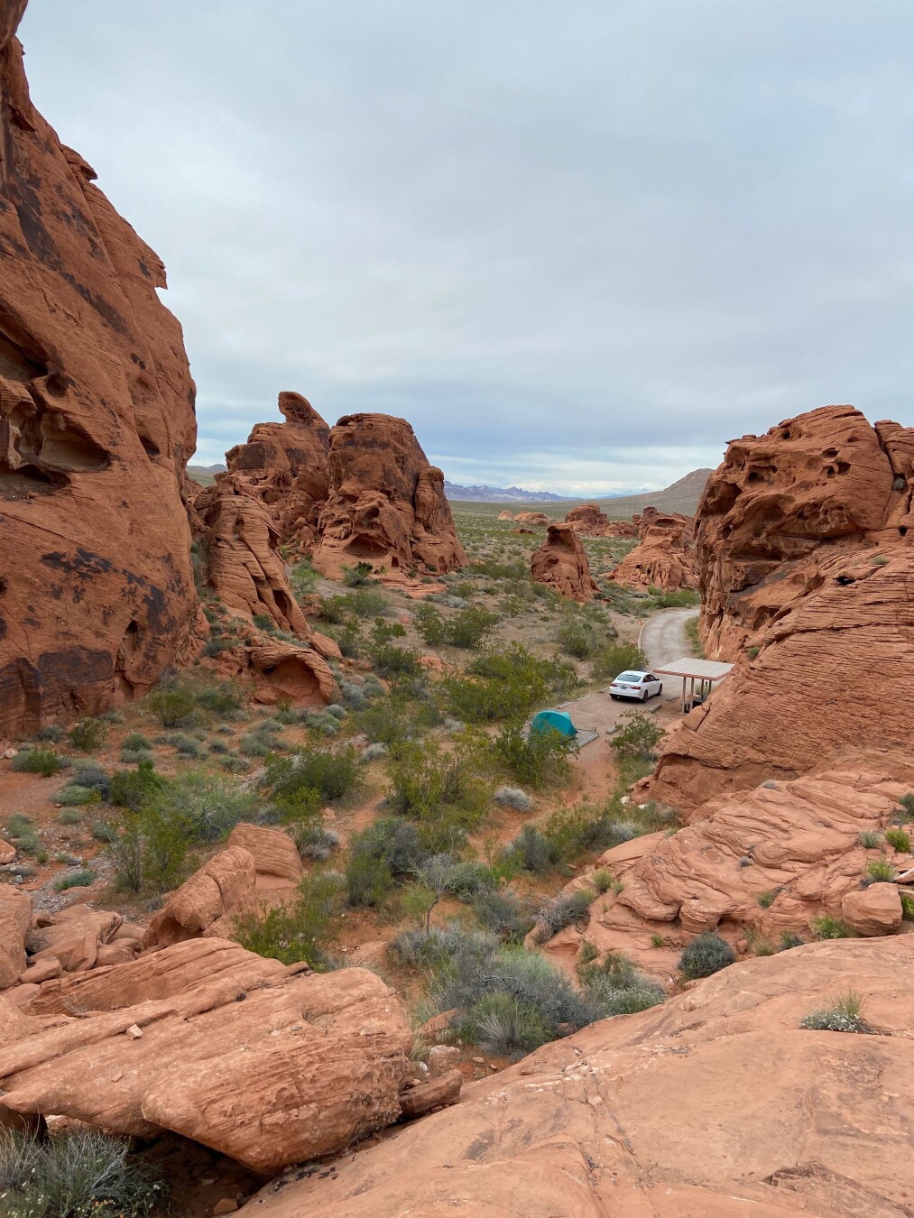 A landscape view shows a rocky terrain with red-orange rocks on both sides, leading to a road in the center of the frame. The road has a white car and a shelter beside it. A turquoise-colored tent is pitched near the road. The ground between the rocks is covered with green bushes and sparse vegetation. The sky is overcast and gray. In the distance, a flat landscape with mountains is visible.