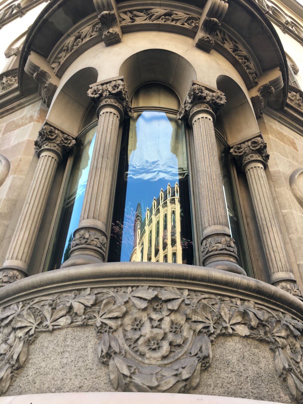 :A low-angle shot of an arched window with ornate stonework. The window is framed by four fluted columns with decorative floral capitals. The lintel above the window features intricate carvings of leaves and flowers. In the reflection of the glass, a building with multiple stories and balconies is visible. The sky is blue and clear. The stonework is a light beige color, and the overall aesthetic of the window is classical and elegant.