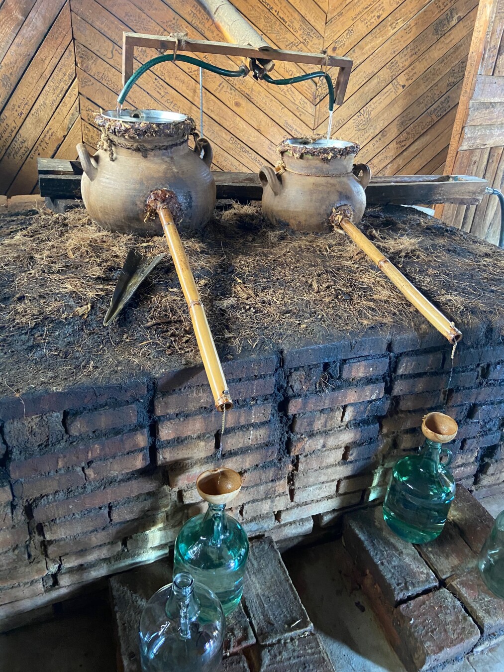 The image shows a rustic distillery setup with two large, round clay pots placed on top of a brick structure covered in dry grass or hay. A green hose-like structure connects the two pots, with water dripping from the pots through bamboo pipes into glass bottles below. Each bottle has a wooden funnel placed on top. The background features a wooden wall with inscriptions