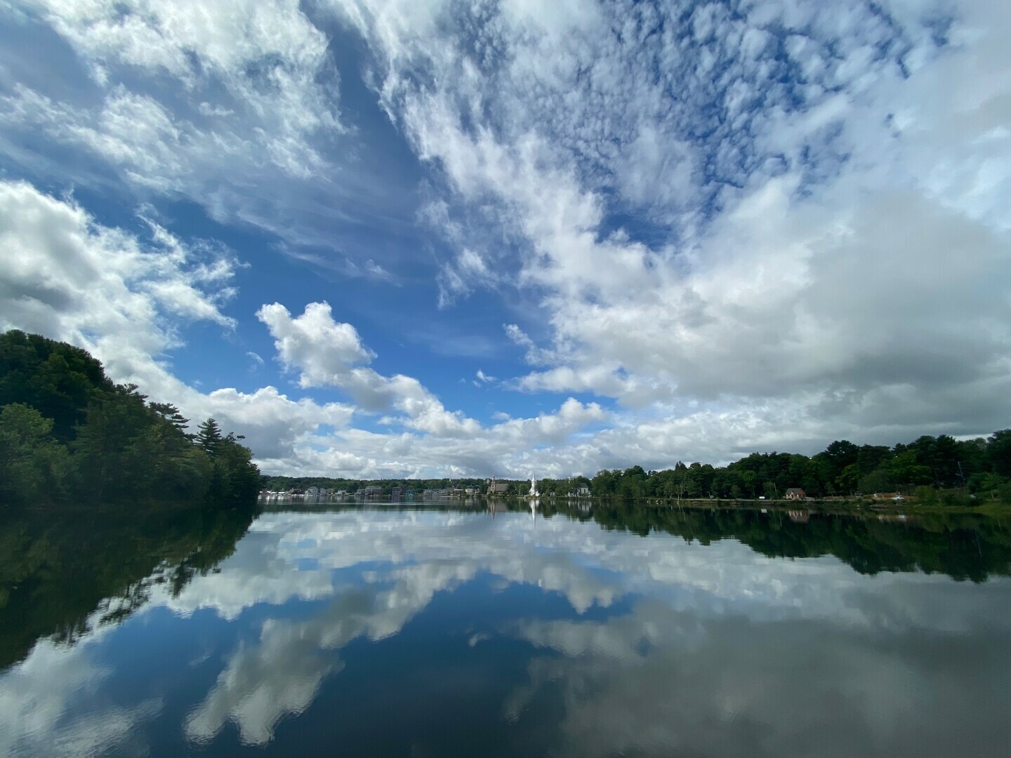 The photograph depicts a serene body of water, reflecting a dynamic sky filled with a mix of blue and white clouds. The water's surface mirrors these clouds, creating a symmetrical effect. On either side of the water, lush green trees line the banks, adding a touch of nature. In the distance, a small town with a church steeple can be seen, completing the picturesque scene.