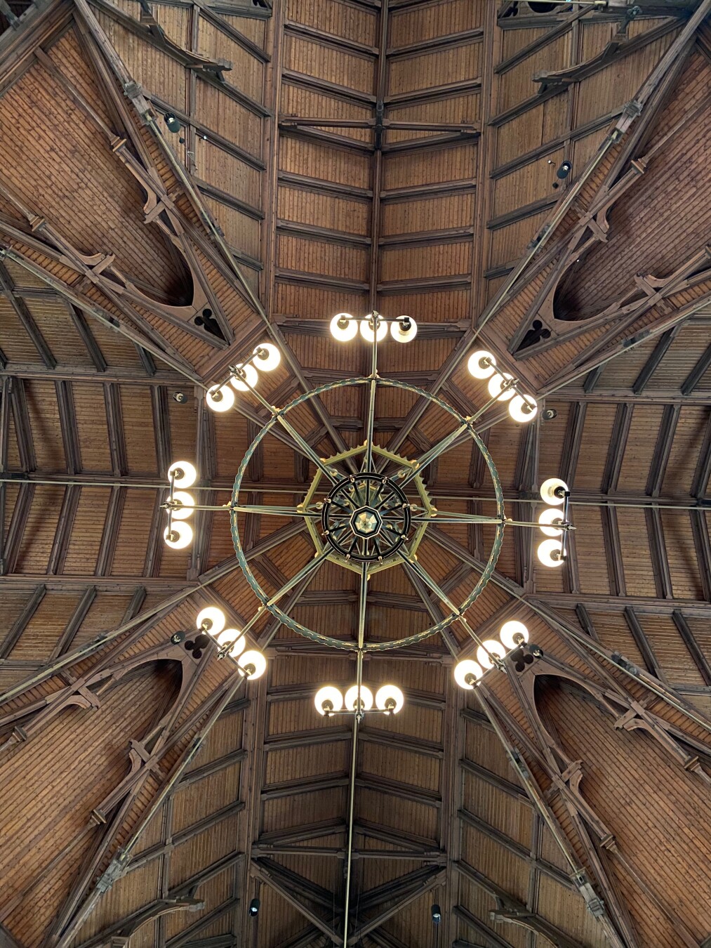 The image shows a view looking straight up at an ornate wooden ceiling with a large chandelier hanging in the center. The ceiling is constructed of wooden planks, and the roof structure is visible, with wooden beams and supports creating a geometric pattern. The chandelier is circular with a decorative center and radiating arms, each holding three light bulbs. The overall aesthetic is vintage and architectural, with a focus on symmetry and detail.