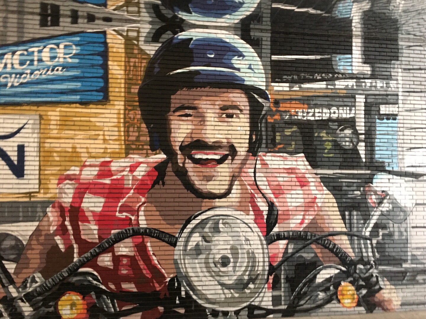 A mural on a brick wall shows a man on a motorcycle smiling. He is wearing a red and white plaid shirt and a blue and black helmet. The motorcycle headlight is round and silver. Behind him are buildings and signs, including one that says 