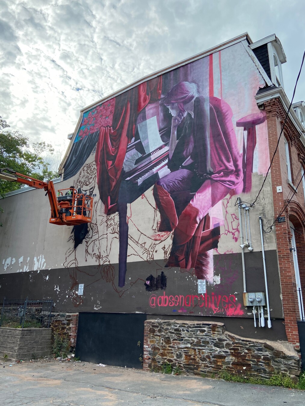 A low-angle shot of a mural being painted on the side of a brick building under a cloudy sky. The mural depicts a woman with her head down playing a piano, with red curtains and a partially drawn tiger behind her. An orange cherry picker is positioned to the left of the mural, with a worker in it. The bottom of the building has a dark lower half and graffiti, including 