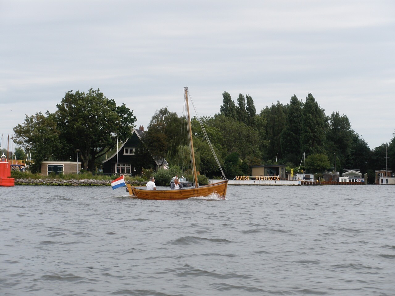 A wooden sailboat with a Dutch flag sails on a river. Two people sit in the boat, and in the background, there are houses and trees. The sky is cloudy. (AI-generated AltText and Hashtags)