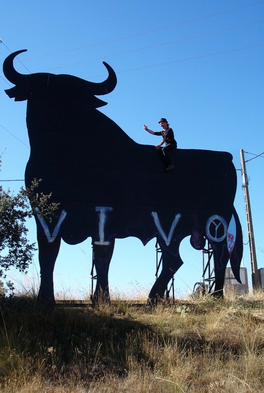 Me riding a black Osborne billboard bull I encountered on one of my hikes in Spain, way back then. It has 'V I V O' sprayed on its legs. I'm waving triumphantly at the photographer. The weather is georgeous and sky is christal blue.
