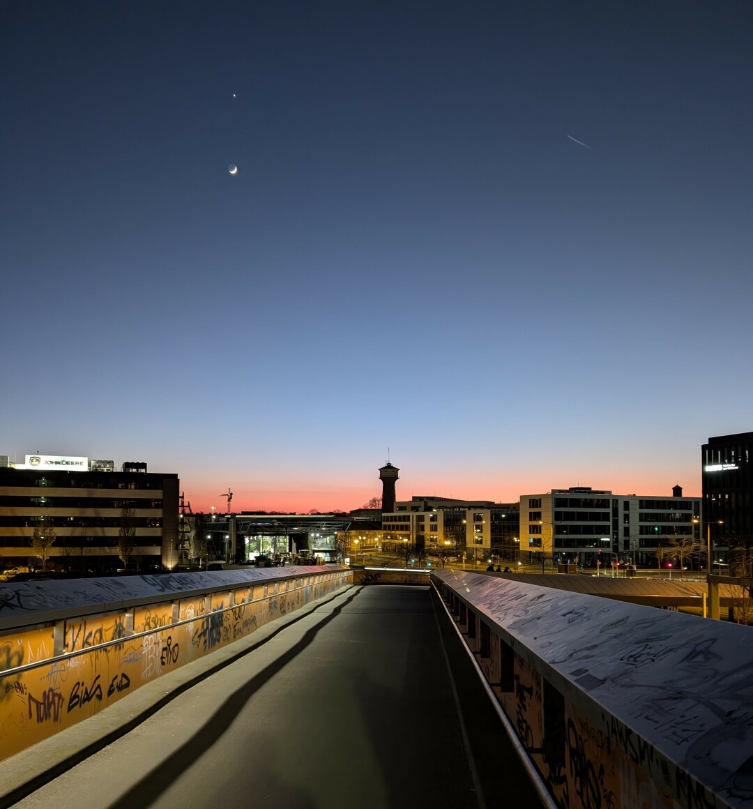 Walking on a bridge in the direction of a sunset above some buildings and a water tower. The moon is visible on the top left side of the picture, above the moon you can see Venus as a bright light.