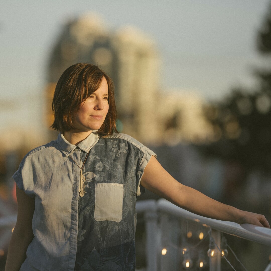 A two-spirit person with light skin and shoulder-length brown hair leans against a railing and links off to the right. She is wearing a blue patchwork sleeveless shirt. Behind he rin soft focus looms a large structure, perhaps a grain silo.