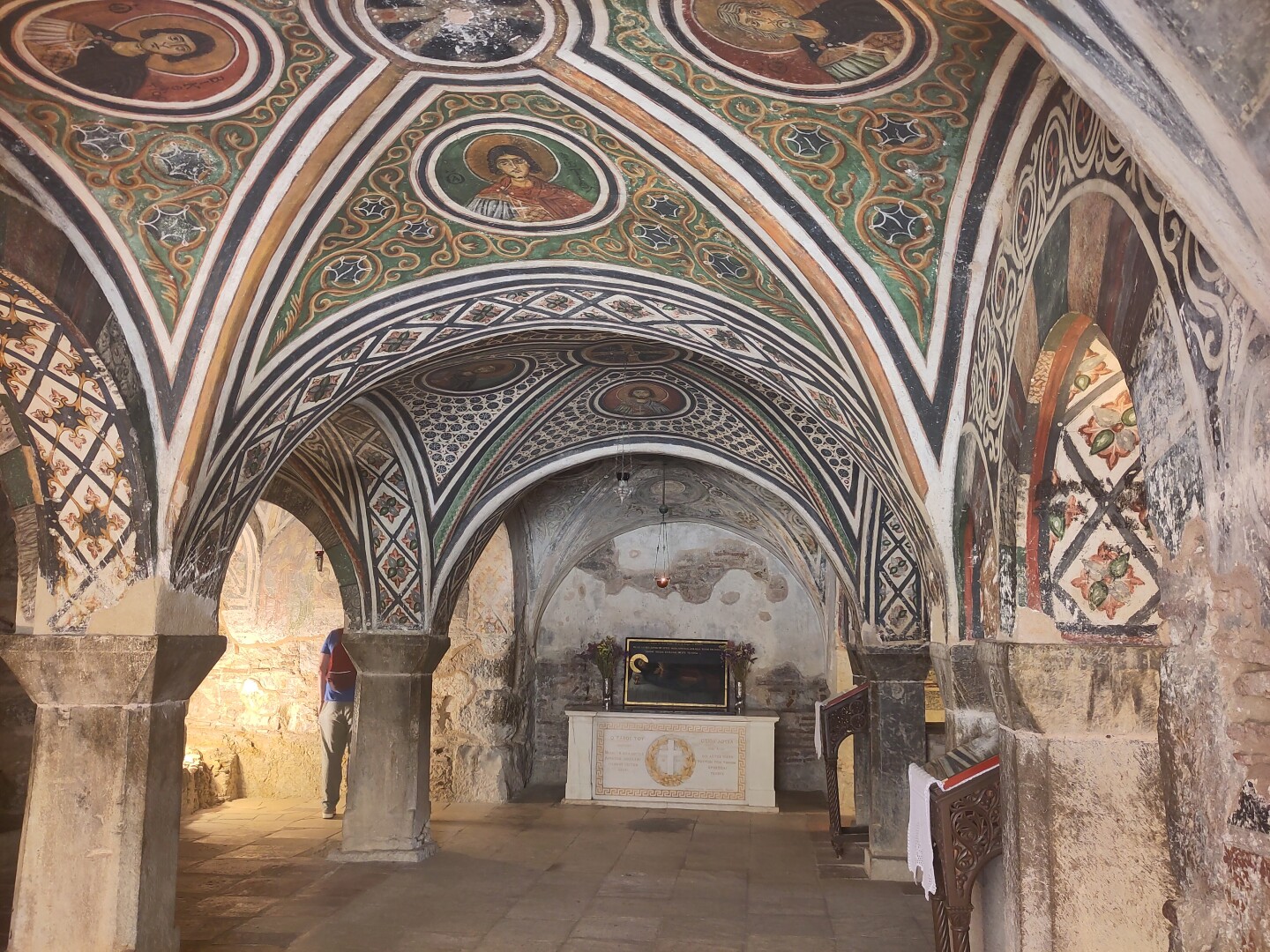 The lower level of the monastery full of richly painted vaulted ceilings, each partition showing an icon of a different saint within colorful geometric designs. A simple white marble altar sits against the far wall.