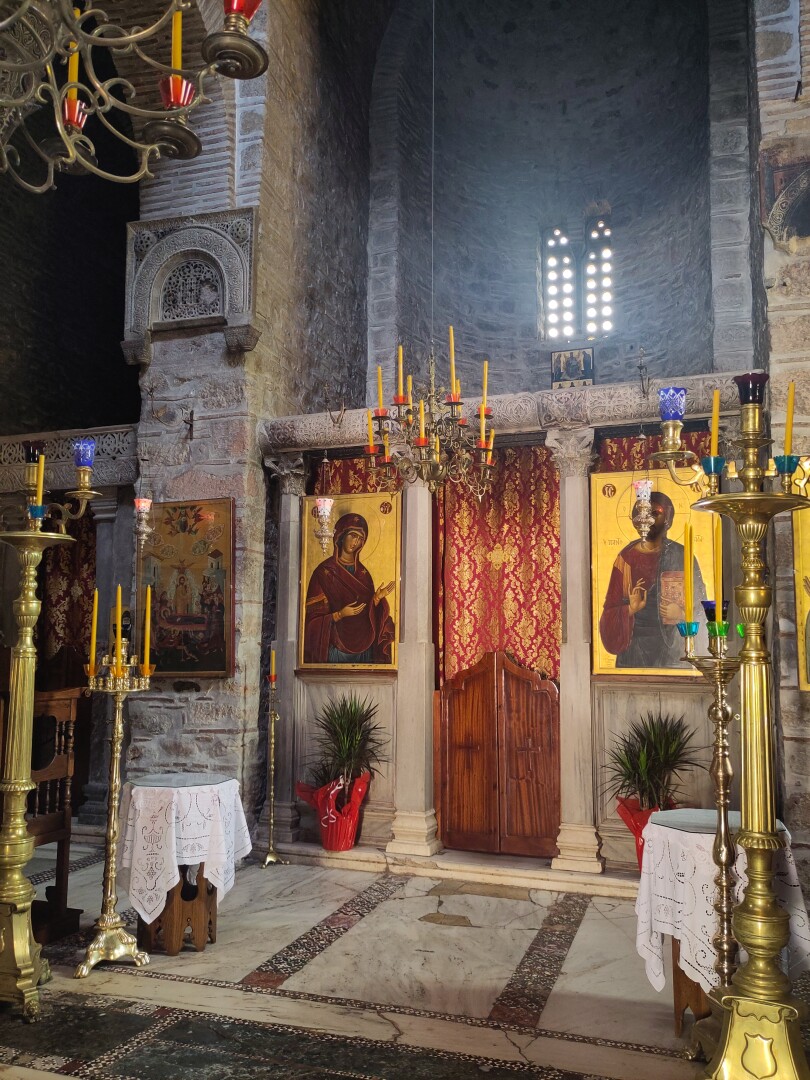 A view of what looks like the confessional: a red-gold curtained off doorway with low wooden doors, flanked by two gold-leafed icons (Mary on the left) and a couple of potted plants. On either side are tall gold candlesticks in front of the church's pillars, another icon hanging on each.
