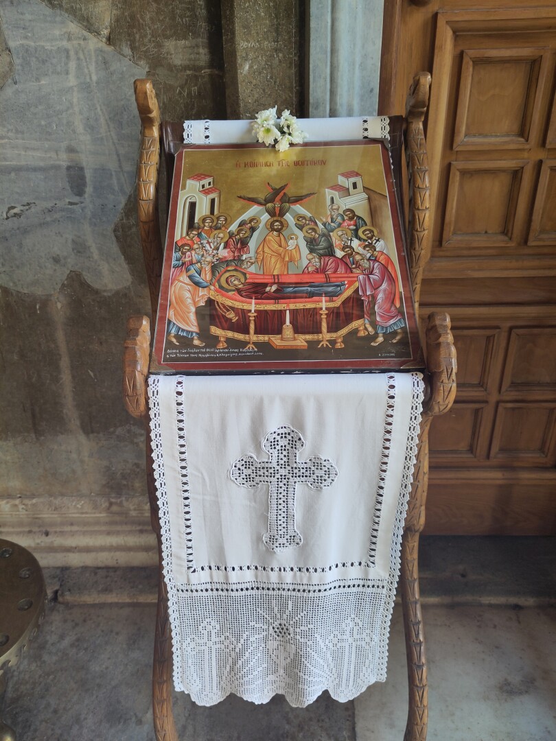 A wooden stand holding aloft a gold-leafed icon, depicting a scene of Mary laying in a red velvet lined coffin, surrounded by a crowd of saints watching over her, a bearded male figure holding a baby in the center (I thought it was Jesus except he'd also be the baby? maybe it's Joseph?) and a multi-winged angel floating above.