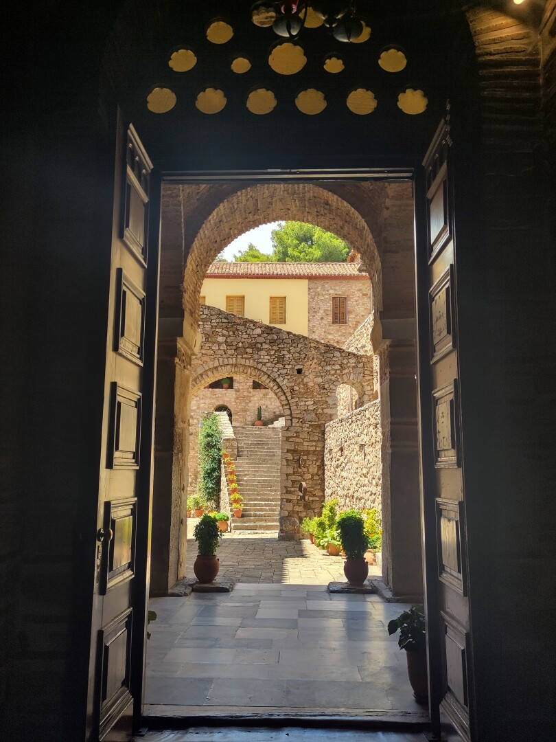 A view through an doorframe topped by a small arch window made of many little circles on top, through a stone arch with potted plants on either side, into an inner courtyard with another stone archway of which the top runs diagonally across, and lastly in the courtyard background, a set of stairs leading up to more buildings.