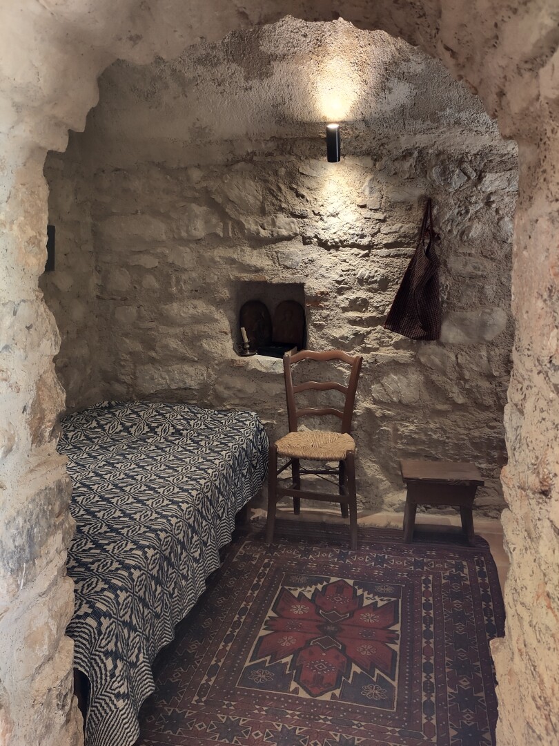 A view through a stone archway of a small room as used by monks in earlier times, decorated with a simple rug, a bed, chair and low stool, with a small cubby hole and some hooks to hang stuff from on the wall. The cubby hole is decorated like a tiny shrine with a couple small icons and candles.