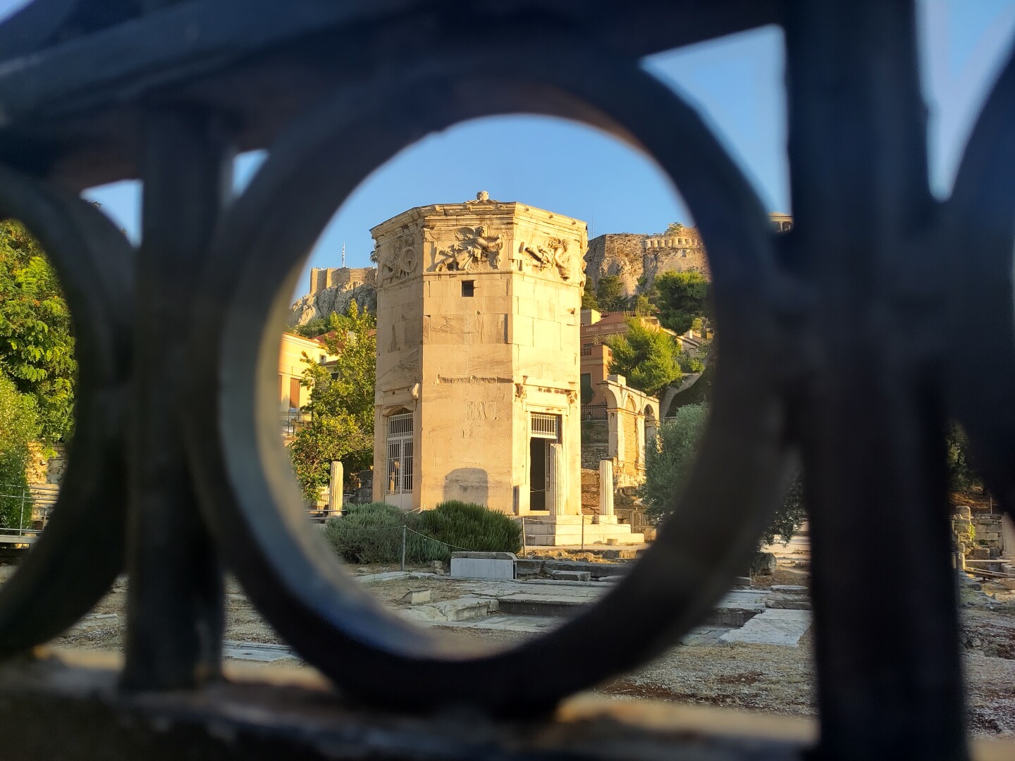 The Tower of the Winds captured in late afternoon sunlight within the natural frame of a fence, shaped like a circle within a square.