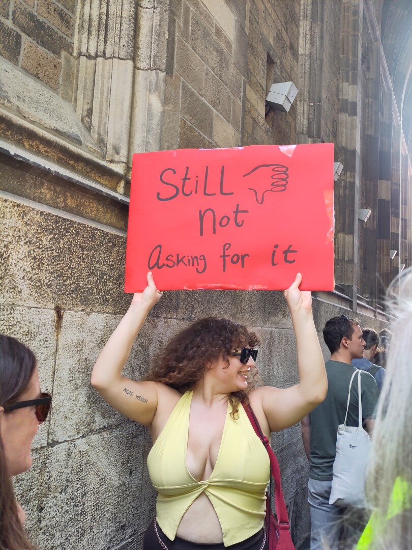 Focus on one woman wearing a small yellow halter top with a split in front showing her stomach, holding up a sign: "Still not asking for it"