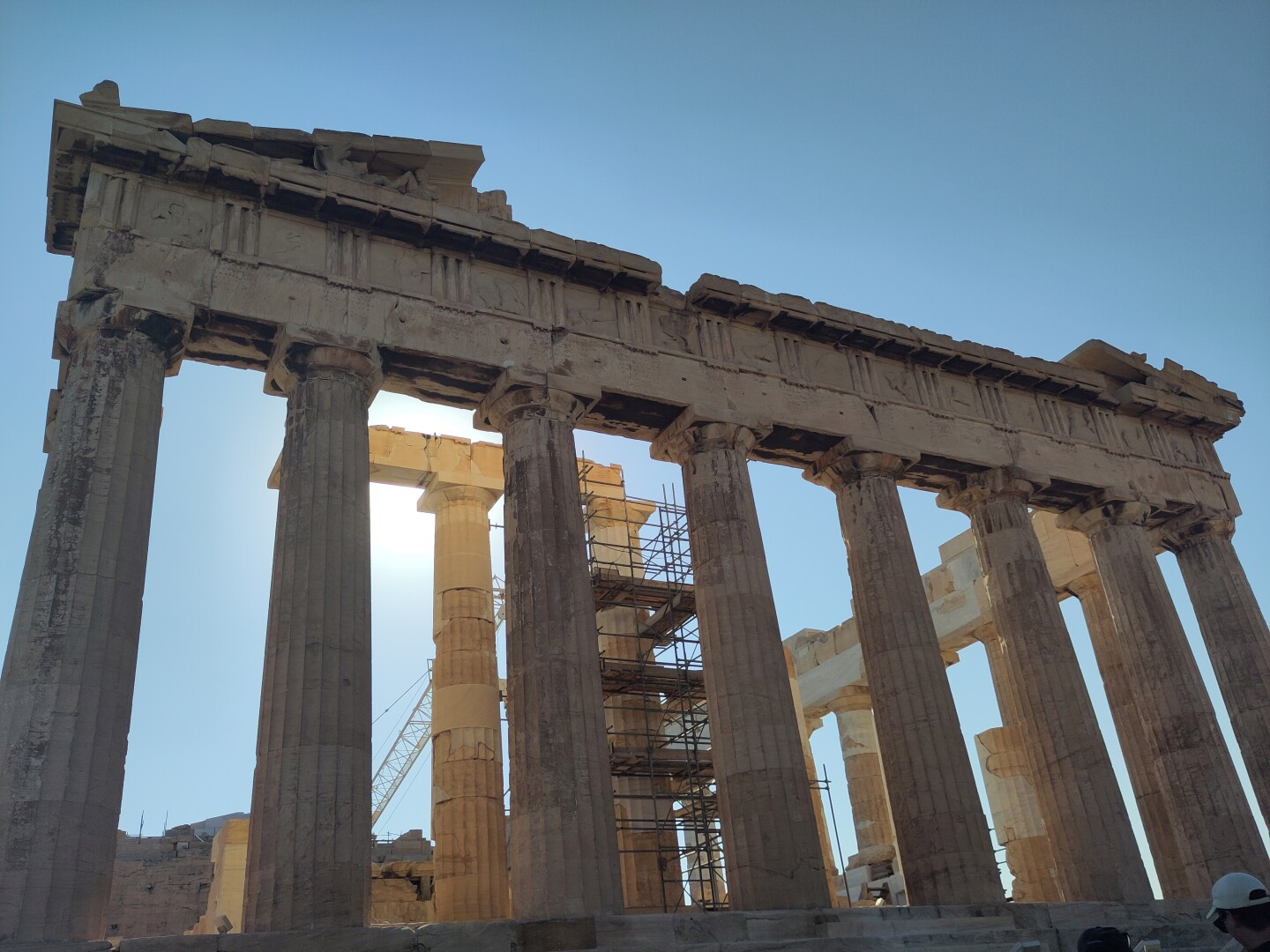 The side facade of the Parthenon, the famous temple dedicated to Athena on the Athenian Acropolis, with eight columns supporting the top. More clear blue skies and the sun shining bright behind it.