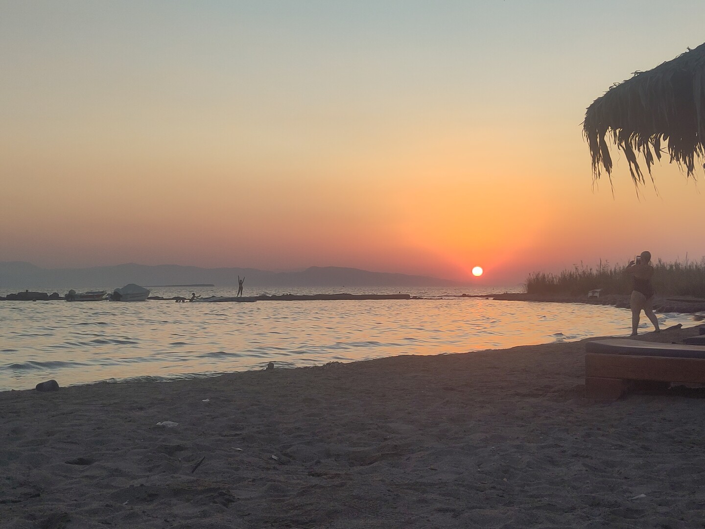 Sunset on the beach, showing the sands up front with the sea behind it, the sky in orange-blue gradients emanating from the sun setting between far away mountains on the horizon.