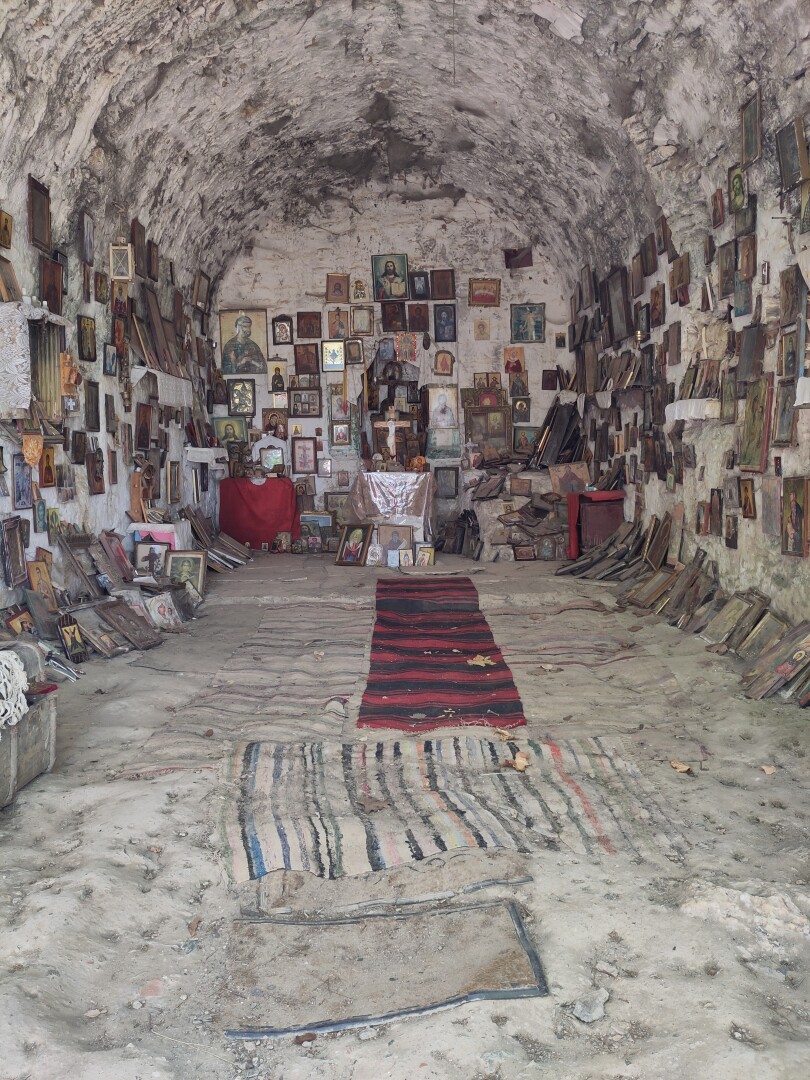 A small cave-like chapel (?) with every wall entirely full of icons of different saints, with a few worn carpets on the stone ground leading up to a small altar with a statue of Jesus on the cross.