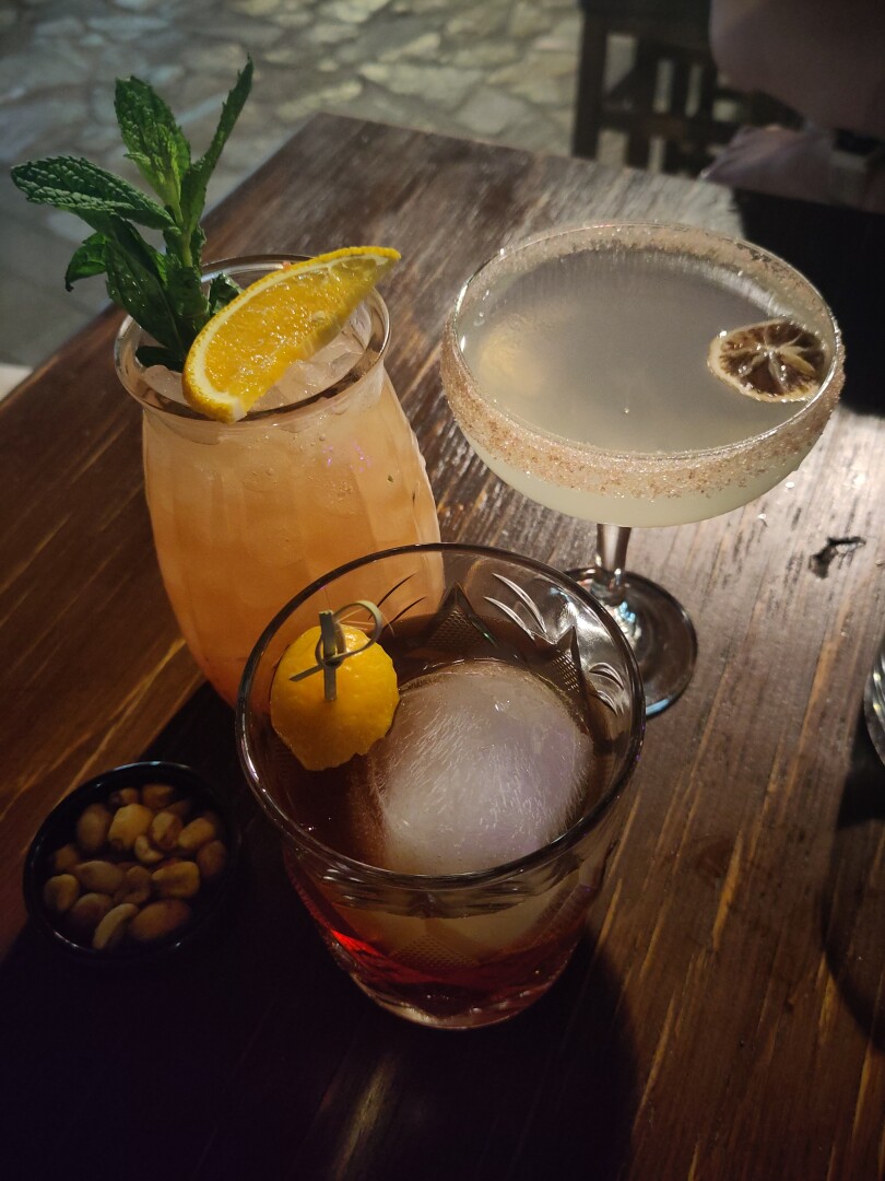 Three cocktails on a wooden table.
On the right is a classic salt-rimmed margarita.
On the left is an orange drink with green leaves and a slice of orange, the Monkey's Tail: scotch whiskey, pineapple juice, orange juice, lime juice, simple syrup, triple sec, blueberry jam.
In the middle is a tumbler glass containing clear amber liquid around an ice ball, Le Stroumpfette: mushroom infused bourbon, maraschino liquor, drambuie, sweet vermouth, agave syrup, bitters.