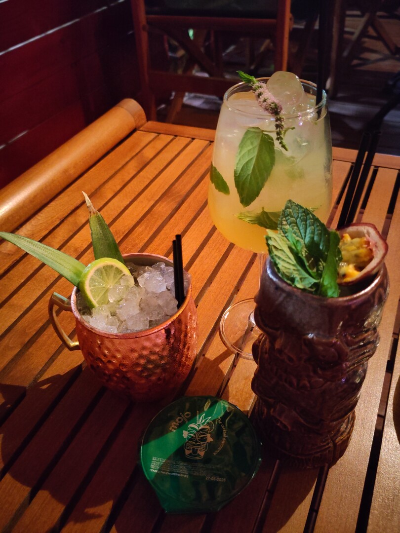 Three cocktail drinks sitting on a wooden table, surrounding a small green container of complimentary nuts with the bar logo "Mojo" on it.
The left drink (Old Cuban) is in a copper mug with ice, green leaves and a slice of lime visible: it contains rum, gomme, lime, spearmint and moscato.
The middle drink (Fly Me To The Moon) is in a wine glass, yellow mixed with mint leaves and ice: it contains gin, maraschino, pear puree, gomme and lime.
The right drink (Zombie) is in a tiki mig with sprigs of mint and half a passion fruit on top: it contains rum, orgeat, cinnamon, passion fruit, lime and bitters.