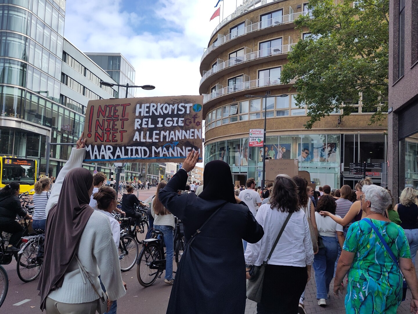 More people from the back, focused on two women wearing hijabs and holding up a sign that says in Dutch:
"Niet herkomst
Niet religie
Niet alle mannen
Maar altijd mannen"

(Translated English:
"Not origin
Not religion
Not all men
But always men")