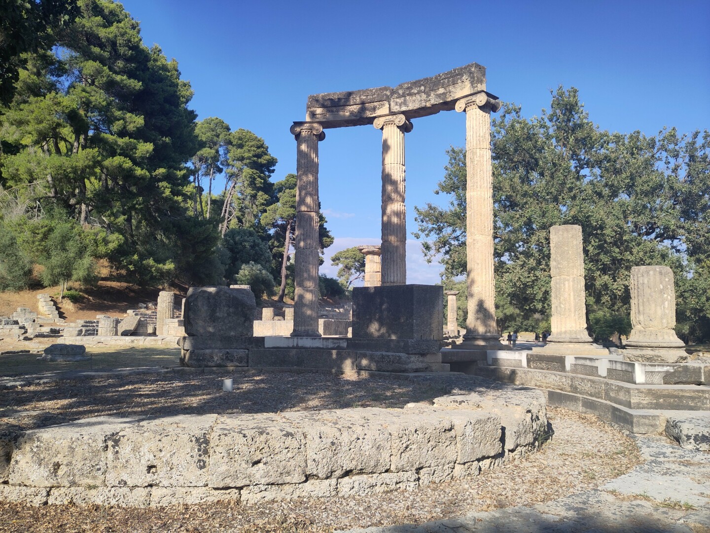 The Phillippeion, the only circular building in Ancient Olympia. There is a circle of low stones of the original walls up front and three pillars still standing tall in the background.