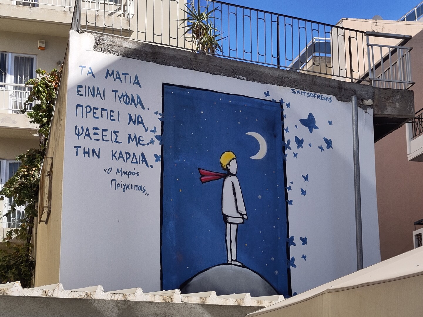 A building wall with a street painting of Antoine de Saint-Exupéry's The Little Prince: the famous picture of him standing on a gray planet, looking towards the moon on the right, against a dark blue sky. There is some Greek text on the left side of the picture, probably a book quote?